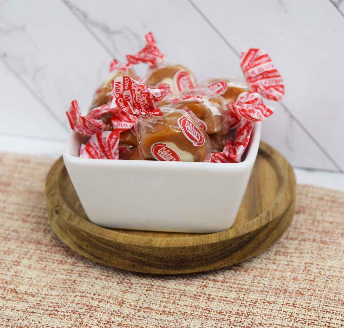 White bowl filled with Goetze's Caramel Cream wrapped candies on a wooden coaster with a woven mat and white wall tile background