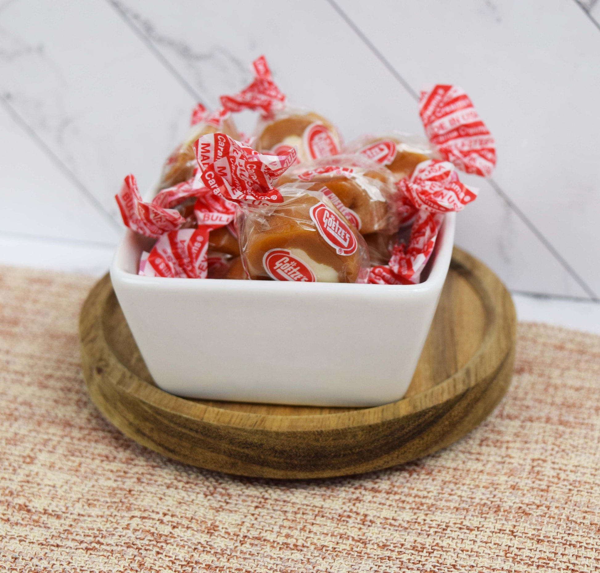 White bowl filled with Goetze's Caramel Cream wrapped candies on a wooden coaster with a woven mat and white wall tile background
